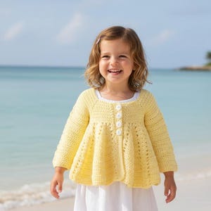 May include: A young child wearing a yellow crochet jacket with white buttons and a white dress stands on a sandy beach. The jacket has long sleeves and a flared bottom. The background features a turquoise ocean and blue sky.