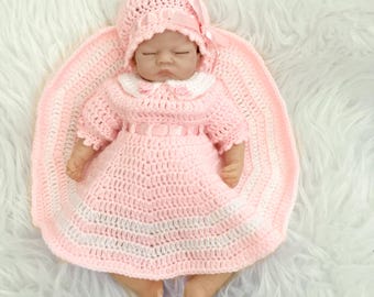 Puede incluir: Una muñeca bebé vestida con un vestido, gorro y manta de ganchillo rosa hechos a mano. El vestido tiene un cuello blanco y un cinturón de lazo rosa. El gorro está adornado con un lazo rosa. La muñeca está sobre una superficie blanca y esponjosa.