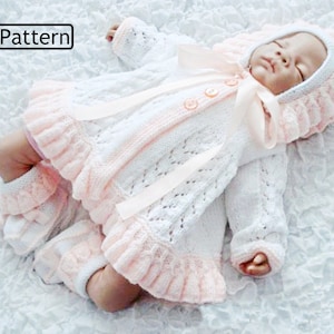 May include: A white and pink knitted baby outfit with a ruffled hem and a matching bonnet. The outfit features a delicate lace pattern and a satin ribbon.