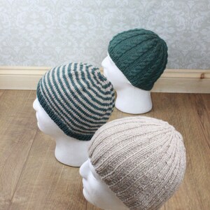 May include: Three knitted hats on white mannequins. The hats are in different styles and colors: one is striped green and beige, one is solid green, and one is solid beige.