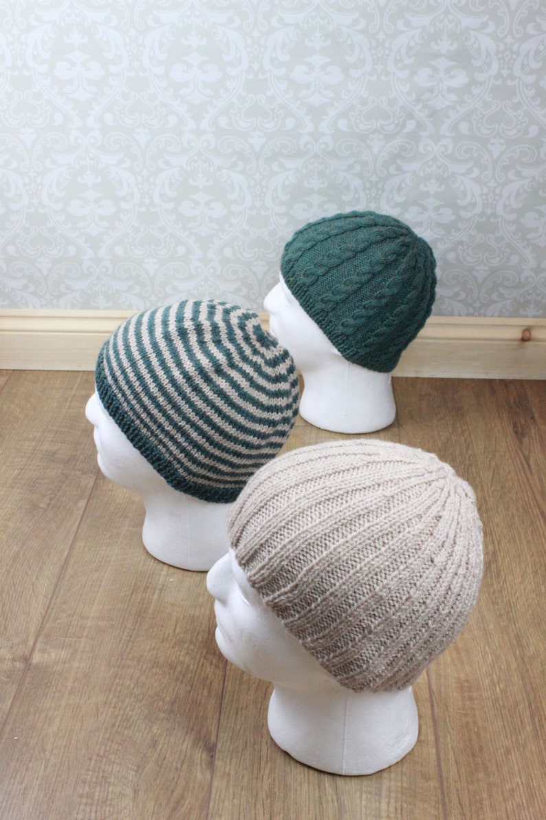 May include: Three knitted hats on white mannequins. The hats are in different styles and colors: one is striped green and beige, one is solid green, and one is solid beige.