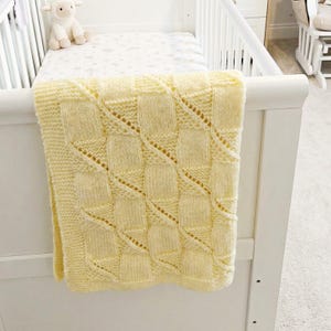 Puede incluir: Una suave manta de bebé de punto amarillo con un patrón geométrico texturizado, colocada sobre una cuna blanca. La manta presenta un diseño repetitivo de cuadrados y líneas diagonales. Un pequeño cordero de peluche está en la cuna.