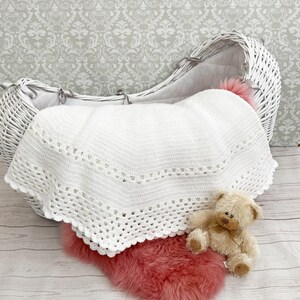 May include: A white, crocheted baby blanket with a scalloped edge is draped over a pink faux fur rug. A white wicker bassinet and a small teddy bear are also in the image. The background features a grey and white patterned wallpaper.