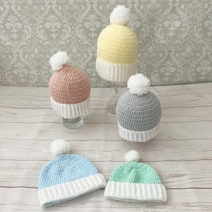 Può includere: Quattro berretti all'uncinetto con pon pon bianchi. I berretti sono di diversi colori: giallo, rosa, grigio, azzurro e verde.