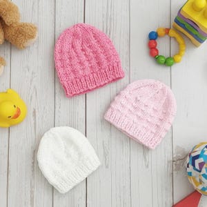 May include: Three knitted baby hats in pink and white are arranged on a light wood-grain surface. The hats have textured patterns. A yellow rubber duck, a toy rattle, and a stuffed bear are also visible.