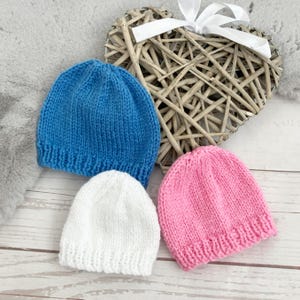 May include: Three knitted baby hats in blue, white, and pink are displayed on a wooden surface. A woven heart-shaped decoration with a white ribbon is in the background. The hats are handmade and appear soft and comfortable.