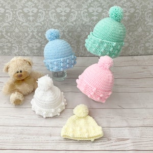 May include: Five crocheted baby hats with pom-poms. The hats are in various colors, including light blue, pink, white, and yellow.