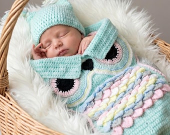 May include: A sleeping baby in a woven basket, wearing a light blue crocheted owl costume with a matching hat. The owl costume features black, pink, and pastel-colored details. The basket is lined with white faux fur.