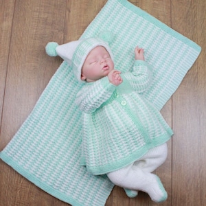 May include: A baby wearing a white and green knitted outfit with a matching hat. The outfit has a ruffled skirt and the baby is lying on a white and green striped blanket.