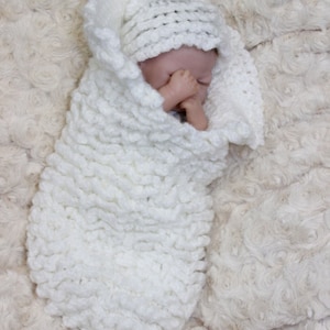 May include: A white crocheted baby blanket and hat set. The blanket is a chunky knit and the hat is a simple stitch. The set is laid out on a white fluffy surface.