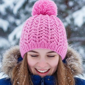 Op de afbeelding: Een roze gebreide muts met een pompon, met een textuurpatroon. De muts is gelabeld met "Shi Fio's Patterns" en "KP579". De persoon draagt een blauwe jas met een met bont gevoerde capuchon, in een besneeuwde buitenomgeving.