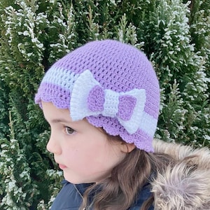 May include: A purple and white crocheted hat with a large white bow. The hat has a scalloped edge and a white stripe.