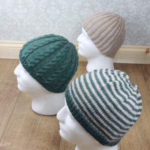May include: Three knitted hats in different patterns: a green cable knit hat, a striped hat with green and beige stripes, and a beige plain knit hat. The hats are displayed on white mannequin heads.
