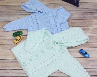 Könnte beinhalten: Zwei gehäkelte Babysweater mit Kapuzen, einer in hellblau und einer in hellgrün, auf einer Holzoberfläche.