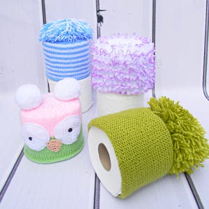 May include: Four toilet paper rolls decorated with knitted hats. One hat is blue and white striped, another is pink with white pom-poms, another is purple and white, and the last is green with a green pom-pom.