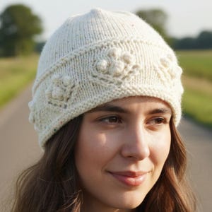 May include: Cream-colored knit beanie with a floral pattern. The hat features a ribbed brim and textured floral designs around the crown. The yarn has flecks of color throughout. A cozy winter accessory.