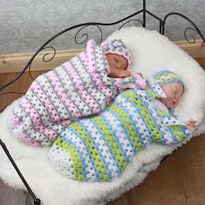 May include: Two crocheted baby sleeping bags, one pink and gray, the other blue, green, and white, with matching crocheted hats. The sleeping bags are laid out on a white fluffy surface.