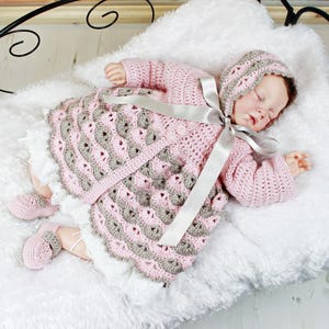 May include: A pink and grey crocheted baby dress with a matching bonnet and booties. The dress has a scalloped hem and a satin ribbon tied around the waist.