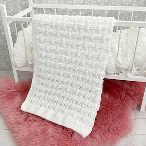 May include: White crocheted baby blanket with a delicate openwork pattern. The blanket is laid out on a pink fluffy rug.
