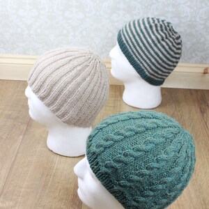 May include: Three knitted hats on white mannequins. The hats are in different styles and colors: a beige ribbed hat, a green cable knit hat, and a striped hat with beige and green stripes.