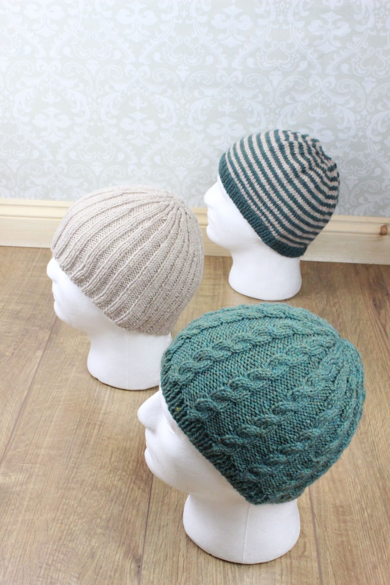 May include: Three knitted hats on white mannequins. The hats are in different styles and colors: a beige ribbed hat, a green cable knit hat, and a striped hat with beige and green stripes.
