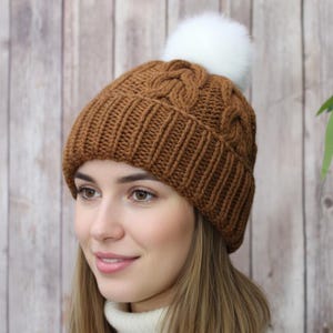 May include: A brown cable-knit beanie with a ribbed brim and a fluffy white pom-pom on top. The hat is made of a soft, warm material, perfect for cold weather. The beanie is a stylish accessory for any outfit.