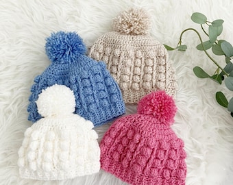 Puede incluir: Cuatro gorros de ganchillo hechos a mano en tonos azul, blanco, beige y rosa, cada uno rematado con un pompón a juego. Los gorros presentan un patrón de puntada en relieve texturizado. Están colocados sobre una superficie blanca y esponjosa.