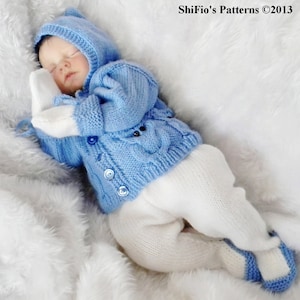 May include: A baby doll wearing a blue knitted cardigan with white buttons and white knitted trousers. The doll is lying on a white fluffy blanket. The text "ShiFio's Patterns ©2013" is visible in the top right corner of the image.