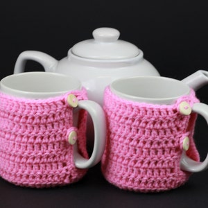 May include: Two white ceramic mugs with pink crocheted cozy covers. The cozies have a button closure and are a great way to keep your hands warm while enjoying a hot beverage.