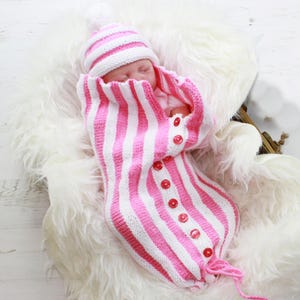 May include: A pink and white striped knitted baby sleeping bag with a matching hat. The sleeping bag has a button closure and a pom-pom on the hat.