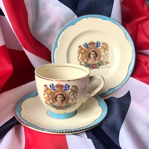 Könnte beinhalten: Ein Vintage-Teetassen- und Untertassen-Set mit einem Porträt von Königin Elisabeth II. Das Set ist mit einem königlichen Wappen und den Worten "Queen Elizabeth II" und "June 2, 1953" verziert. Das Set wird auf einer Union Jack-Flagge präsentiert.