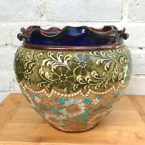Pode incluir: Um vaso grande, redondo e de cerâmica com uma borda festonada. O vaso é decorado com um padrão floral verde e dourado e um esmalte azul na borda.