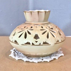 May include: Cream-coloured ceramic vase with a pierced floral design. The vase has a bulbous body and a flared top, accented with gold trim. It sits on a white lace doily, creating a delicate and elegant aesthetic.