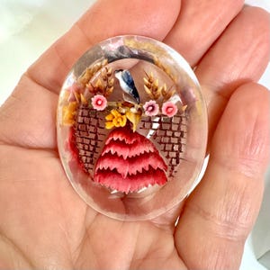 Pode incluir: Um broche oval de resina transparente com elementos botânicos secos incrustados. O design apresenta uma figura central com uma saia de pétalas cor-de-rosa em camadas, pequenas flores amarelas e rosas, espigas de trigo e um pássaro azul.