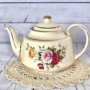 May include: A cream-colored teapot with a gold trim and floral design featuring pink, red, and yellow roses. The teapot is sitting on a crocheted doily.
