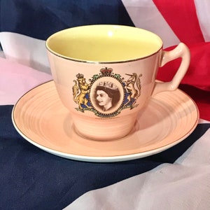 Op de afbeelding: Een roze theekopje en schotel met een gouden rand. Het theekopje heeft een portret van koningin Elizabeth II en het koninklijke wapen. De tekst "June 2nd 1953 Coronation" staat gedrukt op de bodem van het theekopje.