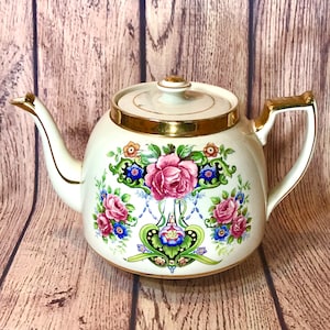 May include: A white teapot with a gold trim and floral design. The teapot has a pink rose, blue and green leaves, and a gold trim around the top.