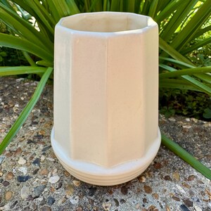 May include: A white ceramic planter with a hexagonal shape and a smooth, slightly textured surface. The planter has a wide, open top and a slightly flared base.