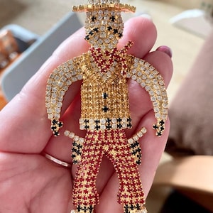 Puede incluir: Un pin de vaquero de pedrería con base dorada. El vaquero lleva una camisa estampada en rojo y negro, pantalones rojos y un sombrero negro y blanco. El pin está cubierto de pedrería transparente, roja y negra.
