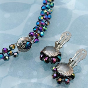 May include: A set of iridescent purple and blue glass bead jewelry, including a necklace and a pair of clip-on earrings. The necklace has a silver clasp and the earrings have silver backs.