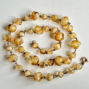 May include: A long necklace made of brown and white glass beads. The beads are faceted and have a swirl pattern. The necklace is strung on a gold chain.