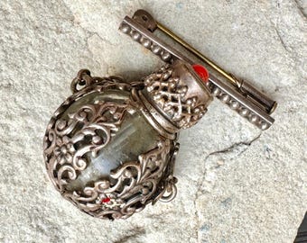 May include: A silver filigree bottle with a red glass stopper and a silver chain. The bottle is empty and has a clear glass interior. The bottle is attached to a silver pin.