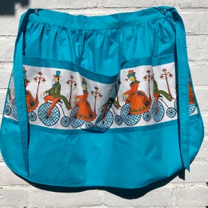May include: A turquoise blue apron with a white band featuring a repeating pattern of people riding bicycles. The people are dressed in vintage clothing and the bicycles have large front wheels.