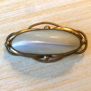 May include: An antique oval brooch with a pearlescent centre and a gold-toned metal frame. The brooch has an intricate design with curved lines and small spherical accents, creating an elegant and classic appearance.