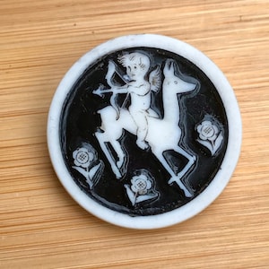 Puede incluir: Un botón de cerámica blanca con fondo negro que presenta un querubín blanco montando un ciervo blanco con arco y flecha. El botón está rodeado de tres flores blancas con centros negros.