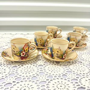 May include: Set of six vintage teacups and saucers featuring a cottage and floral design. The cups and saucers are white with a gold trim. The design depicts a cottage with a thatched roof, flowers, and a blue sky.