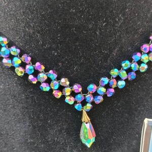 May include: A multi-strand iridescent necklace with a teardrop pendant. The necklace is made of small, faceted beads that are strung together on a black cord. The pendant is a single, large, faceted bead that is suspended from the necklace by a small, gold-colored chain.