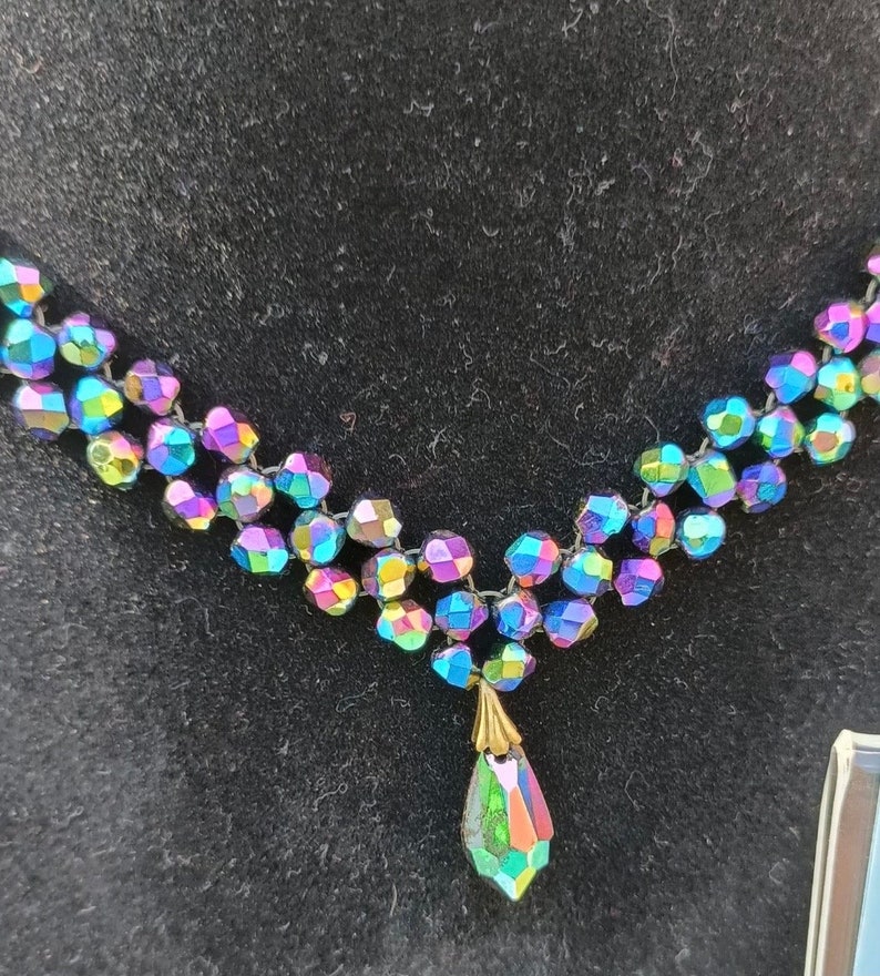 May include: A multi-strand iridescent necklace with a teardrop pendant. The necklace is made of small, faceted beads that are strung together on a black cord. The pendant is a single, large, faceted bead that is suspended from the necklace by a small, gold-colored chain.