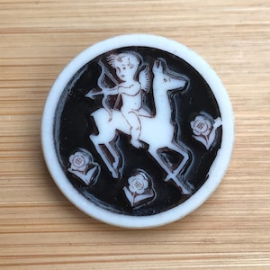 Puede incluir: Un botón de cerámica blanca con fondo negro que presenta un querubín blanco montando un ciervo blanco con arco y flecha. El botón está decorado con tres flores blancas con centros marrones.