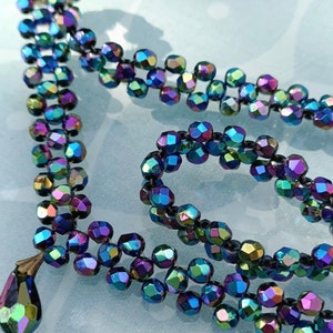 May include: A multi-strand necklace made of iridescent, faceted glass beads. The beads are dark blue, purple, and green, and they are strung on a black thread. The necklace has a teardrop-shaped pendant at the end.